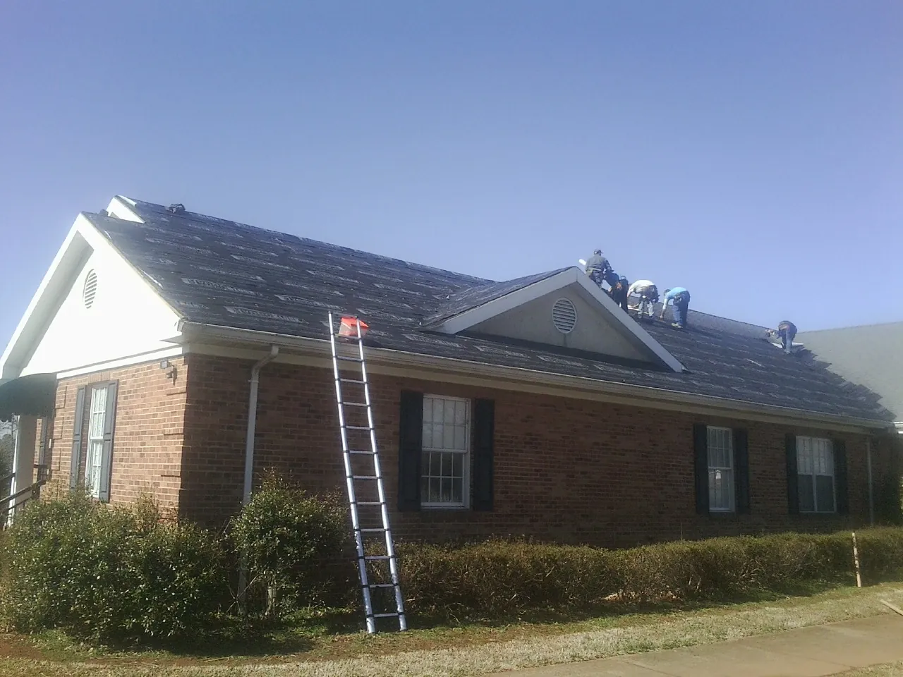 Crew tearing shingles off a brick ranch roof with a ladder set — Metal Roof Repair work by our crew in Peterman