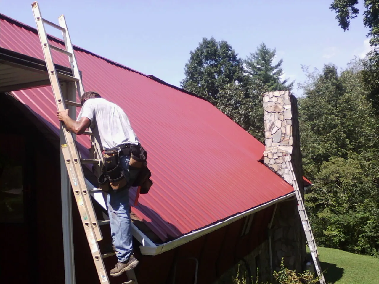 Roofer on a ladder beside a red metal roof and stone chimney — Soffit & Fascia Repair by Roofing Brothers in Peterman